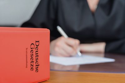 Concept,Court,,Law,And,Law,Firm:,Female,Judge,In,Robe Concept court, law and law firm: Female judge in robe with a German law book in the foreground in her office writing. The label reads: “German laws - text collection”. Selective focus